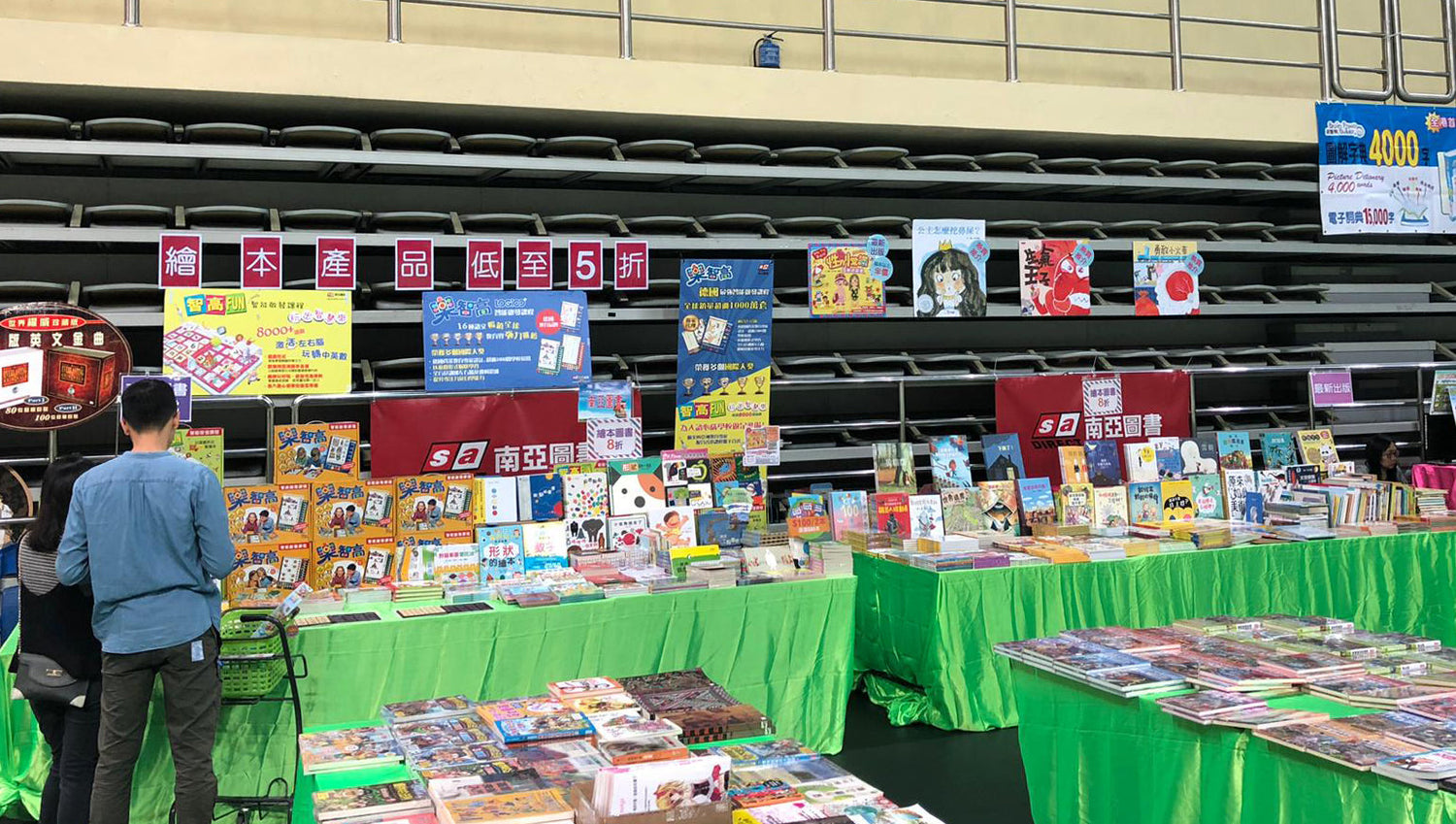 Children's educational books and picture books displayed at a book fair booth with colorful signs and two people browsing.