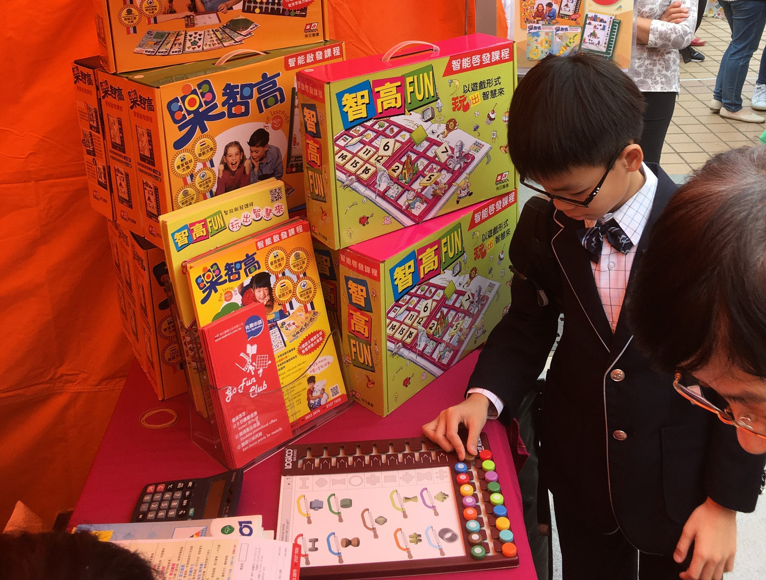 Child in suit interacts with logic puzzle game at educational toy booth, colorful box displays.