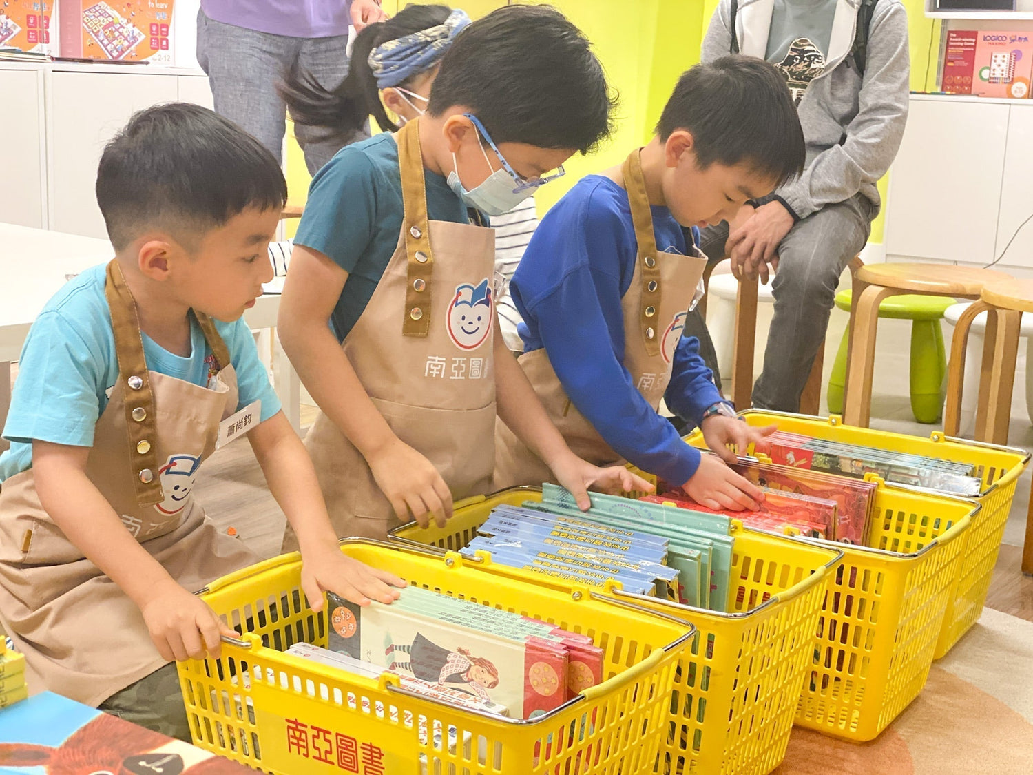 Children in aprons browsing colorful picture books in yellow baskets at a bookstore.