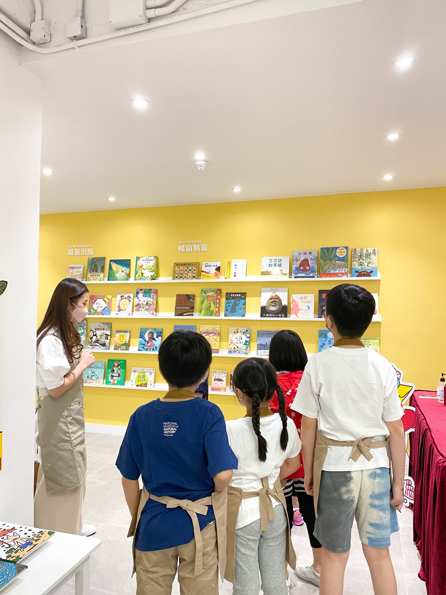 Children in aprons listening to a woman in front of a colorful children's book display.