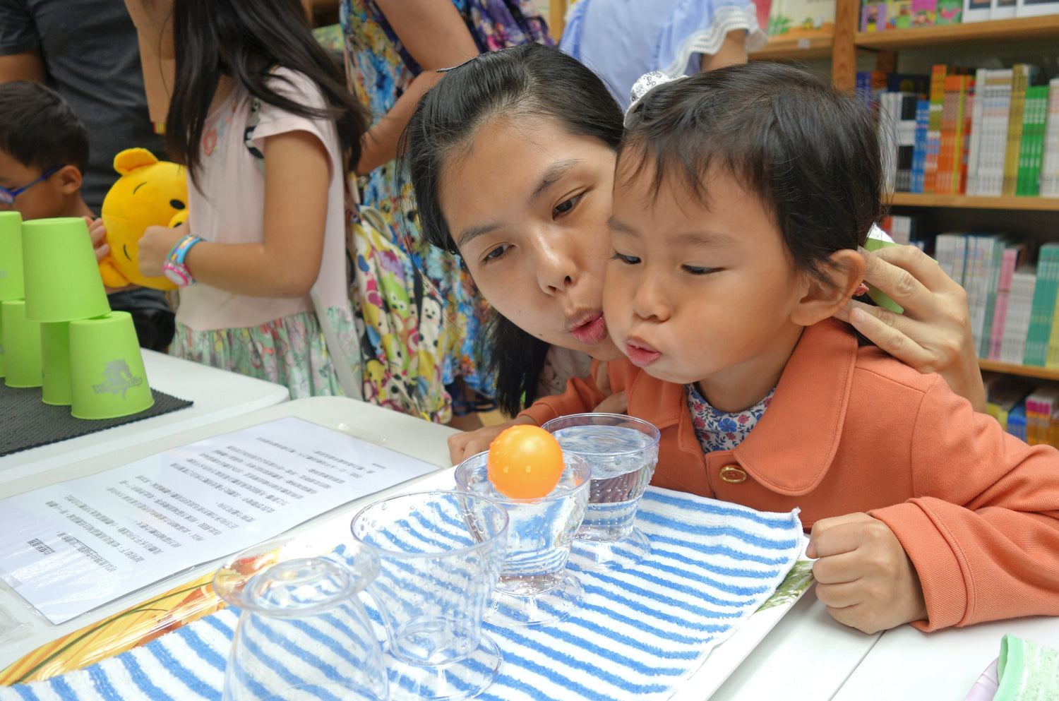 Mother and child blowing ping pong ball over a cup of water at interactive children's book event