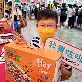 一位戴著黃色口罩的小男孩在書展興奮地舉著「我買咗啦」的牌子和智高FUN課程。