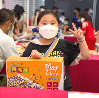 一位戴著白色口罩的小女孩，在書展攤位前抱著智高FUN課程並做出勝利手勢。