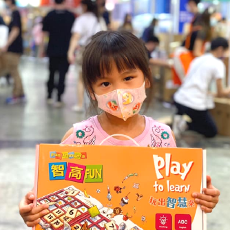 一位戴著粉色口罩的小女孩，在書展上高興地抱著她選購的智高FUN課程。