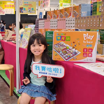 一位小女孩開心地坐在智高FUN攤位前，舉著「我買咗啦」的牌子。